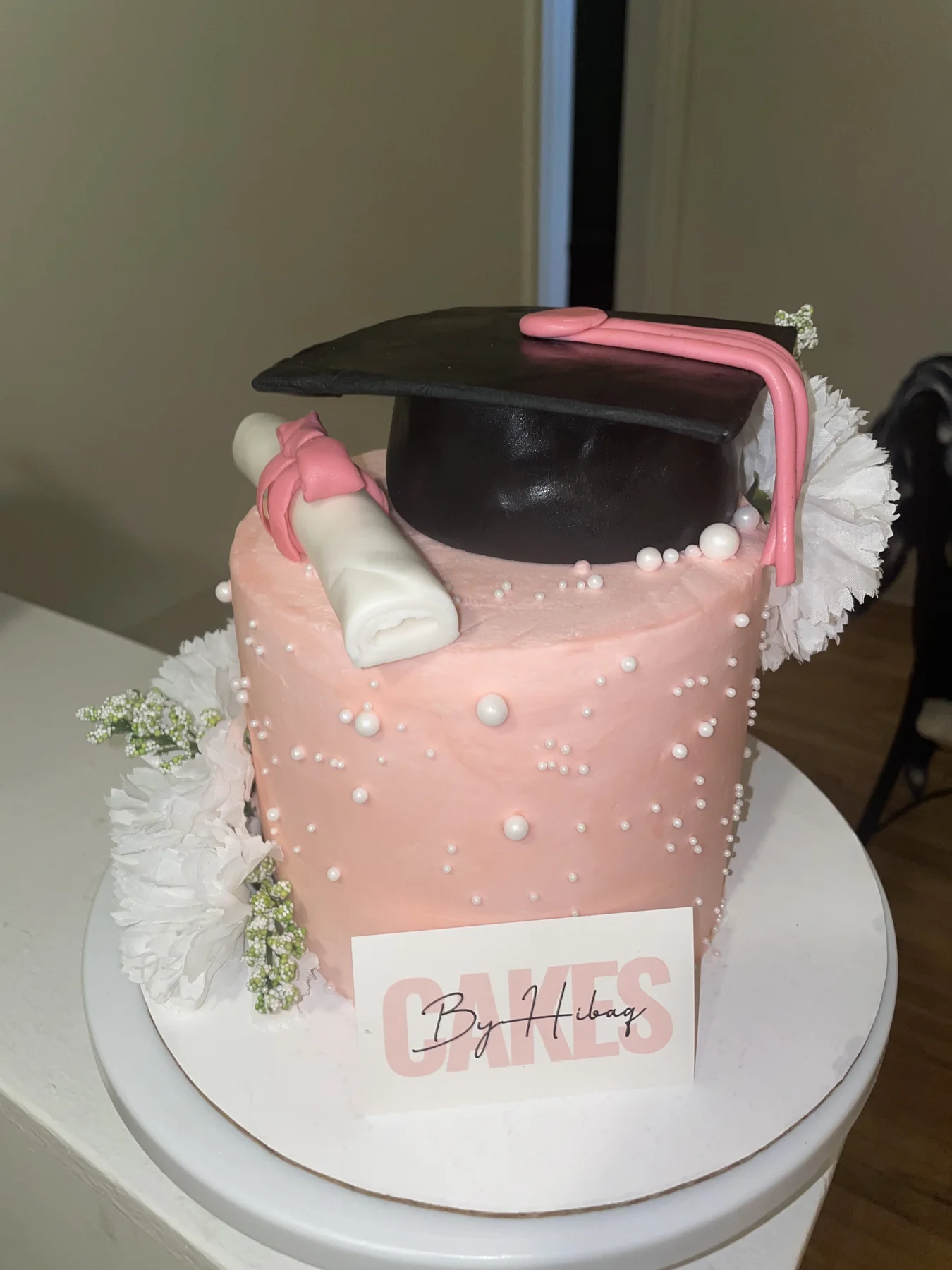 Pink graduation cake with a cap, diploma, pearls, and white flowers